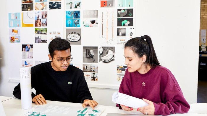 Two students sat working together at a desk. They are both looking at a paper chart and artwork can be seen on a wall behind them.