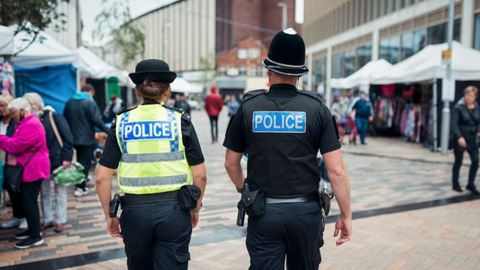 Two police officers walking side by side up a shopping high street. Members of the public can be seen nearby.