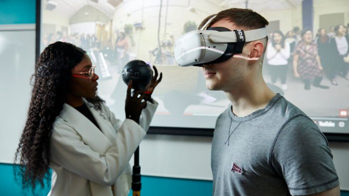 A student stood wearing a VR headset. Next to them another student is holding a circular camera device that's attached to a pole.