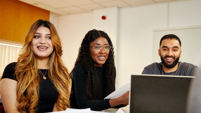 Three students sat at a table working collaboratively. One of them has a laptop open and the other two are holding printed work.