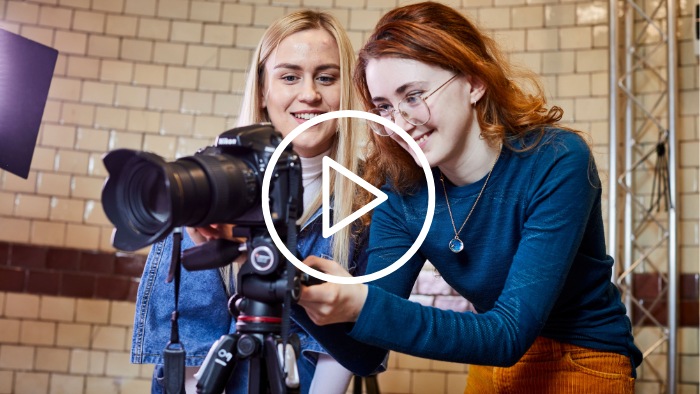 An image showing two students stood setting up a camera shoot and looking into the viewfinder. A large play icon is overlayed on the image.