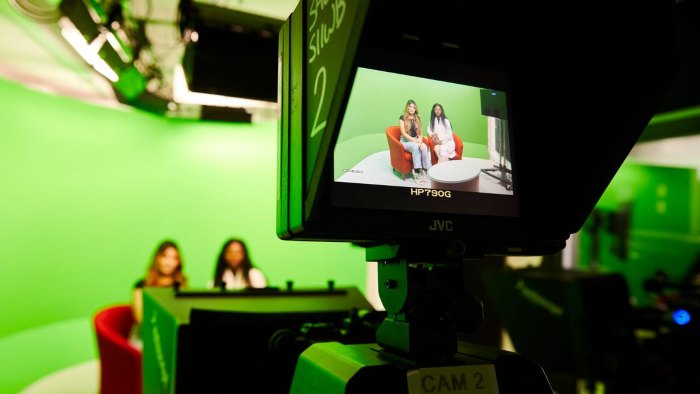 A television camera filming two students sat presenting in front of a large green screen. Both of them can be seen through the camera lense.