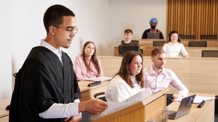 A law student wearing a gown talking to attendees in a mock court room