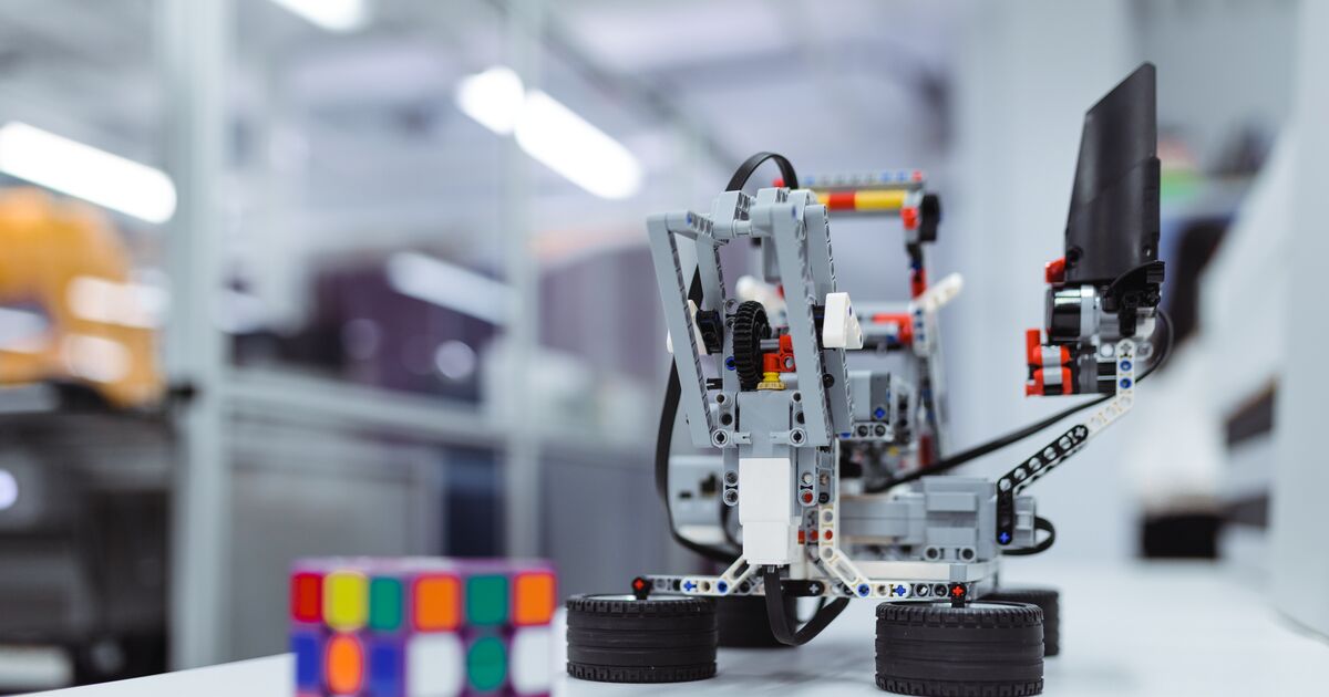 A Lego model robot sat on a table next to a rubiks cube.