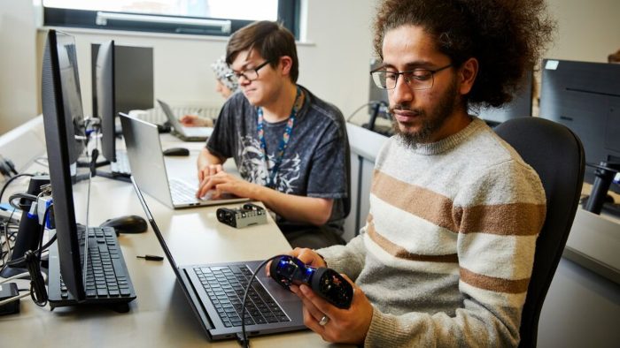 Two students sat at a long desk in front of laptops. One of them is holding a video game controller.