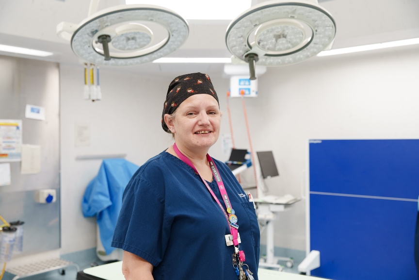 Medical professional in blue scrubs and a patterned scrub cap stands smiling in a surgical room. Behind them are large surgical lights and various medical equipment. They wear an ID badge around their neck.