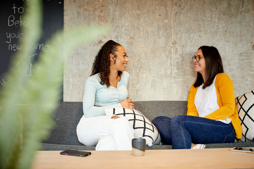 Two students sit on sofa with plant in foreground