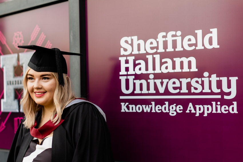 Graduate smiling on graduation day in front of Sheffield Hallam University logo