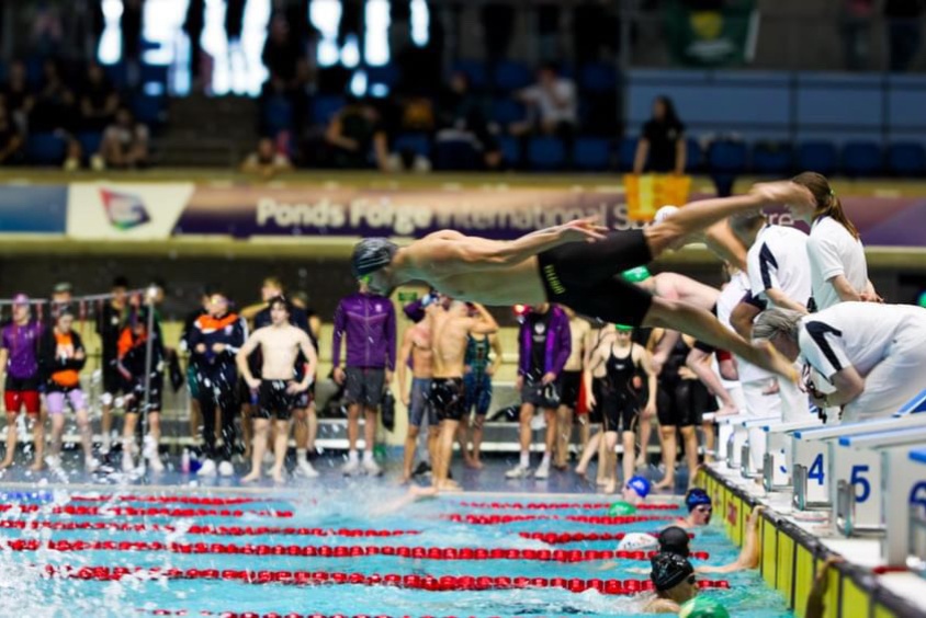 It shows a swimmer in mid-air, diving into the pool with excellent form, arms extended and legs straight. 