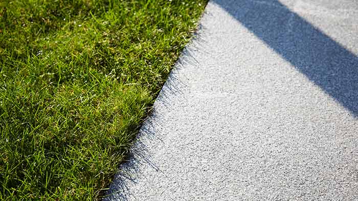 Close-up of the edge of a patch of grass, showing where it meets a pale grey path. The sun is out, throwing shadows on the path.