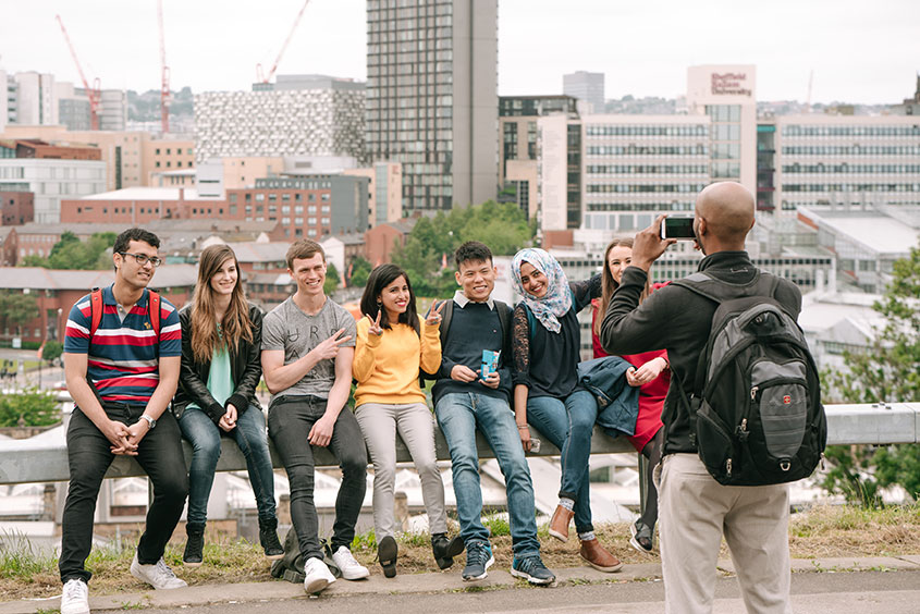 A group of international students sat on a barrier having their photo taken with a backdrop of Sheffield city centre