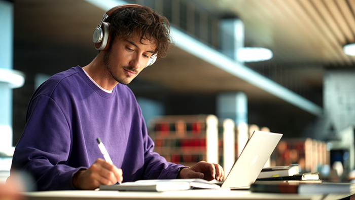 A student working alone in a library. They are male, and wearing headphones.