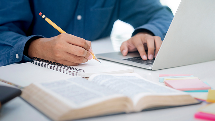 A student making notes as they work on a laptop, a large hard-backed book open on the desk beside them.