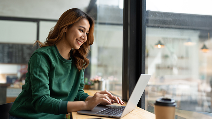 A student smiling at their laptop, by a window in a café.