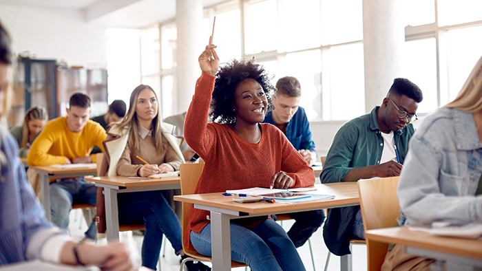 A happy student in a classroom, hand in the air to confidently answer a question. 