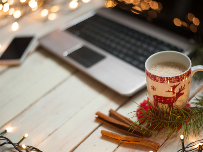 An open laptop sits by a cup of hot chocolate in a Christmas-themed mug, surrounded by fairy lights.