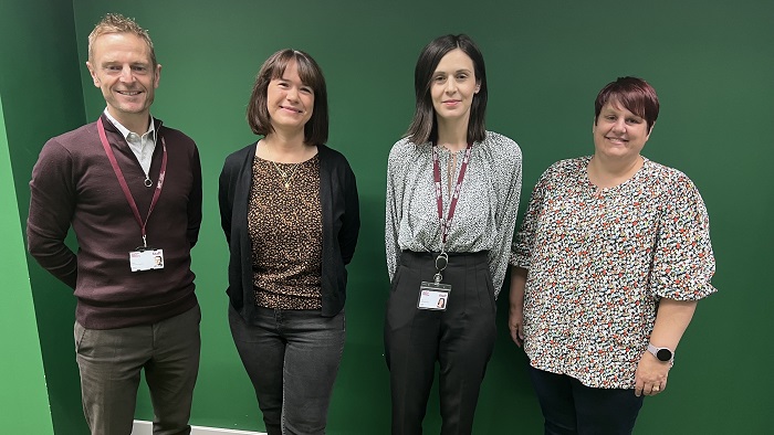 One man and three women stood against a green wall smiling at the camera