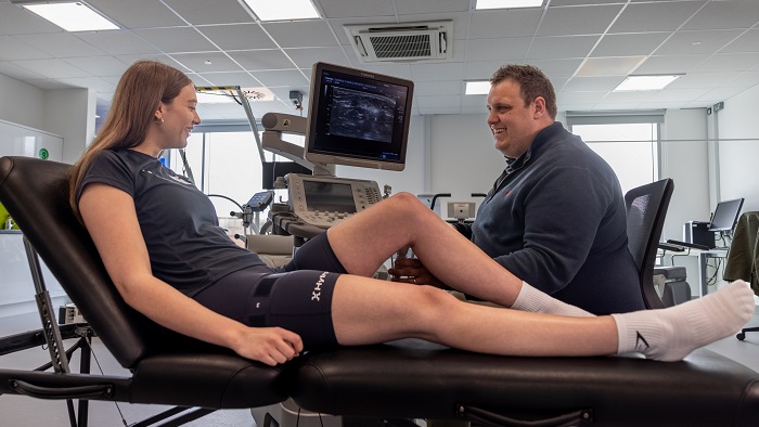 Woman in sportswear sat down chatting to male researcher with clinical machinery in background