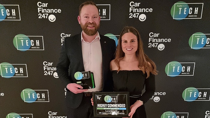 Man in a black suit and woman in a black dress holding awards in front of a banner with logos on