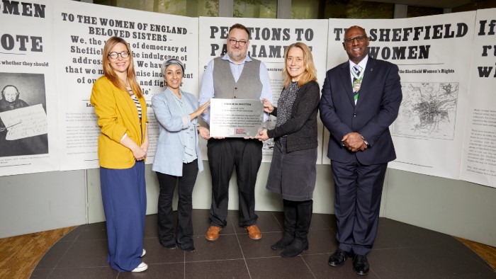 Louisa Harrison-Walker, Abtisam Mohamed, Prof Matthew Roberts, Prof Liz Mossop and Philip Rodrigo stood holding a plaque in front of an exhibition about Sheffield Women's Rights Association. 