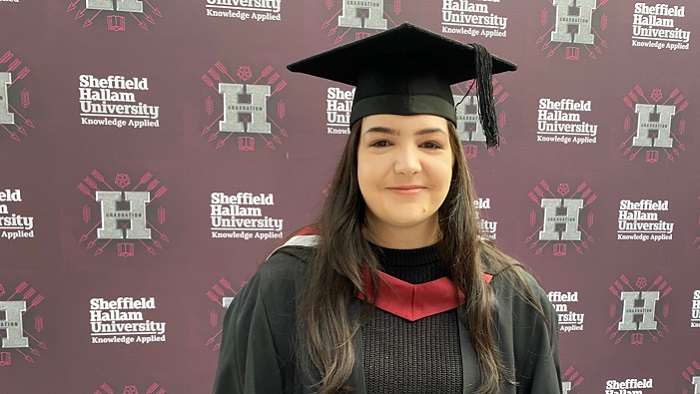 Woman in graduation robes and mortar board in front of a Sheffield Hallam branded background