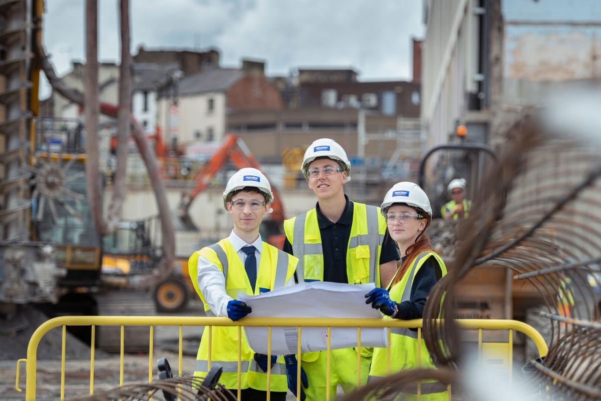 three students wearing hard hats and hi vis on site. They are all holding a site plan.