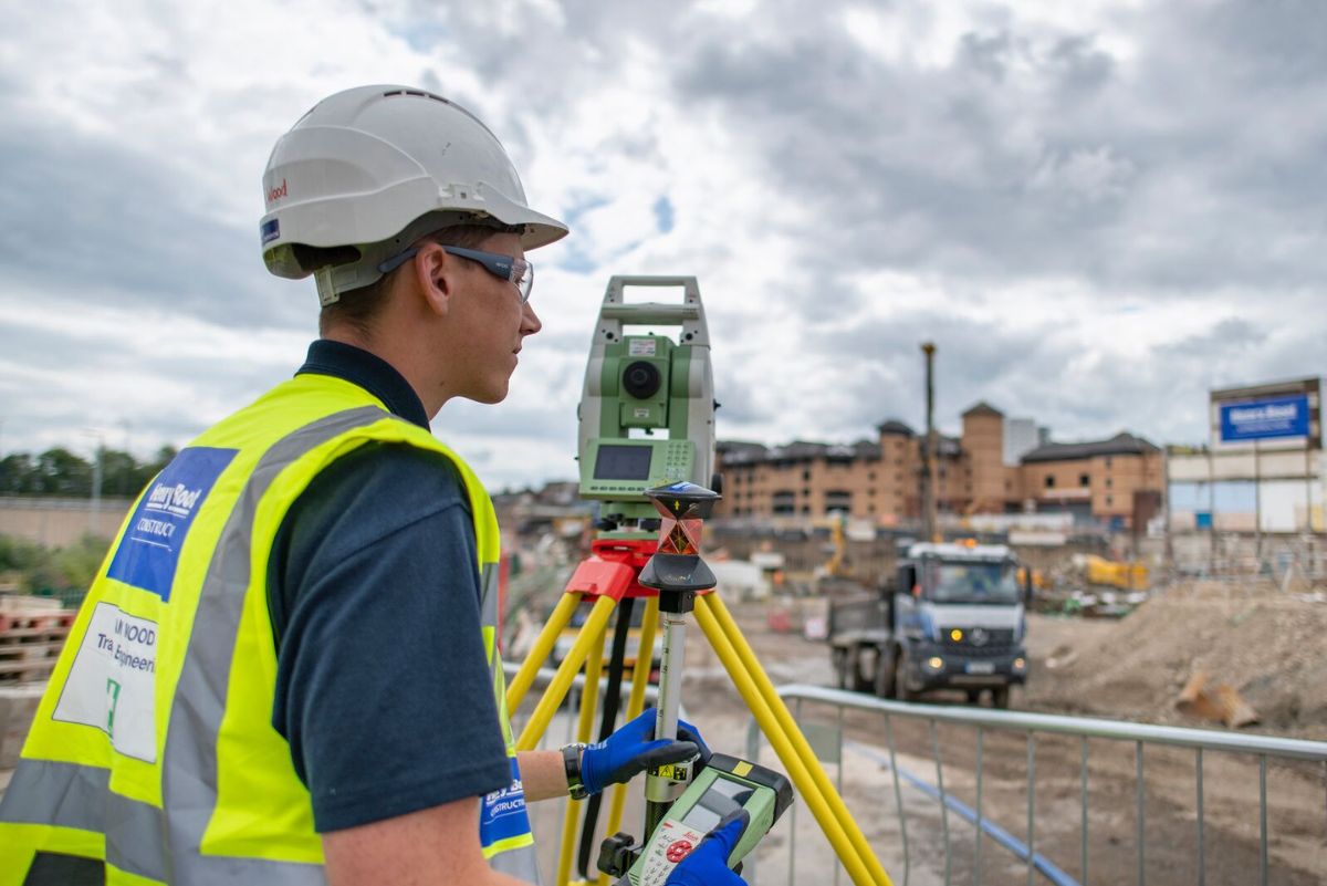 A student wearing a hard hat and hi vis jacket using a robotic total station on site