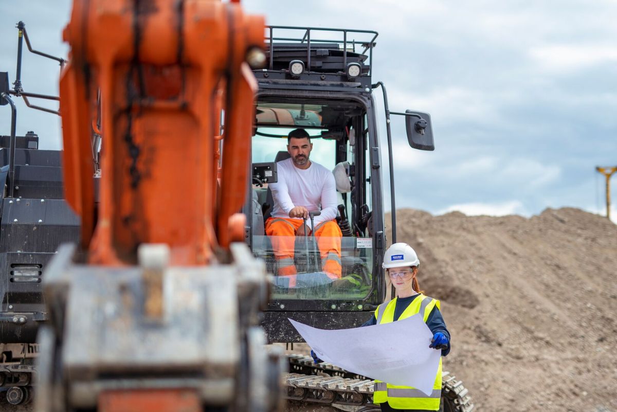 A student on site looking at a plan alongside a man sitting in an excavator