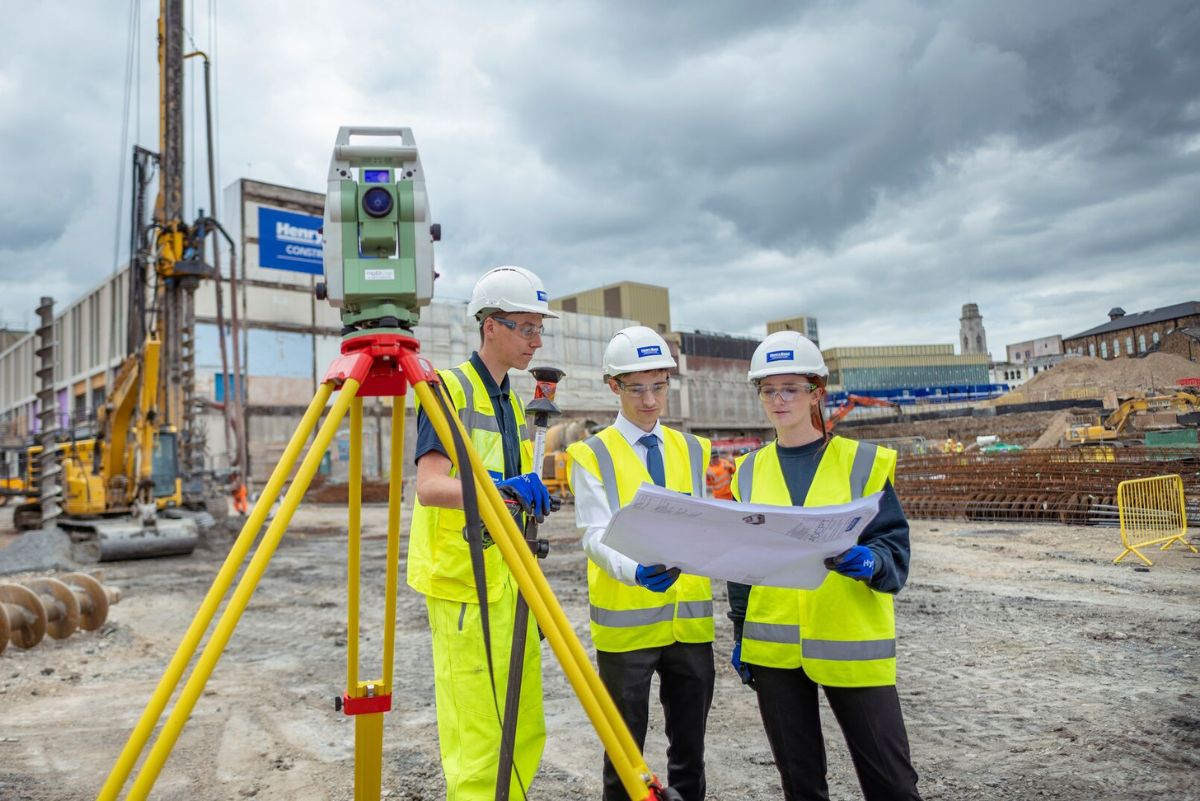 Three people standing in a building site. They are all wearing hi vis and hard hats. They are all looking at a large paper plan.