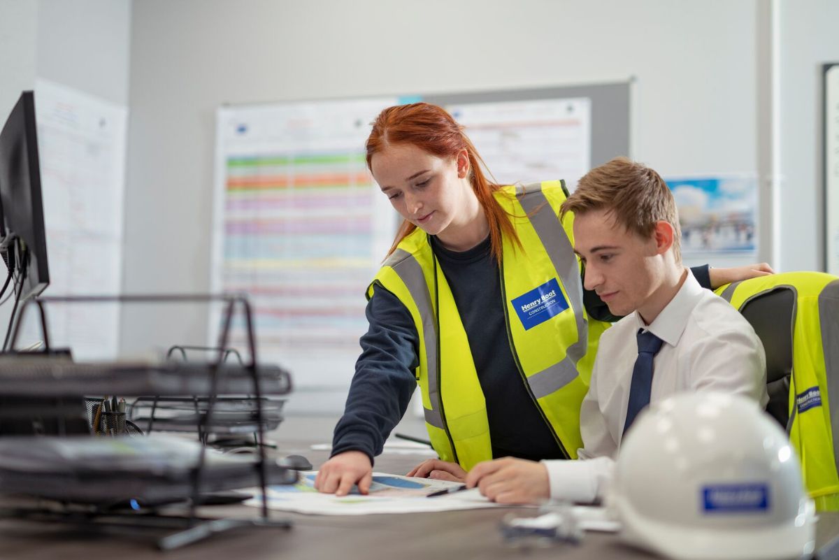 Two people in an office. One person is wearing hi vis and pointing at a document. A person is sat at a desk and looking at the document. 