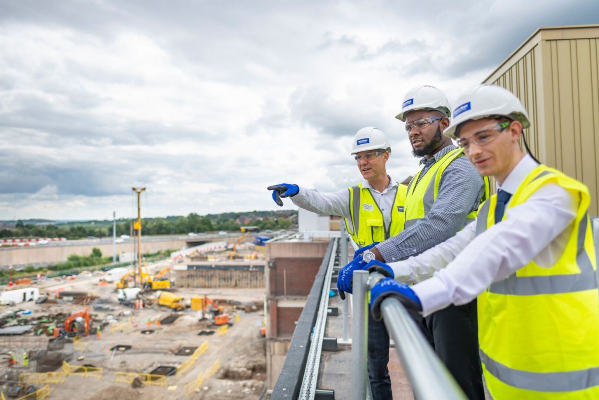 Three people standing on a raised balcony surveying a site. They are wearing hi vis and hard hats. 