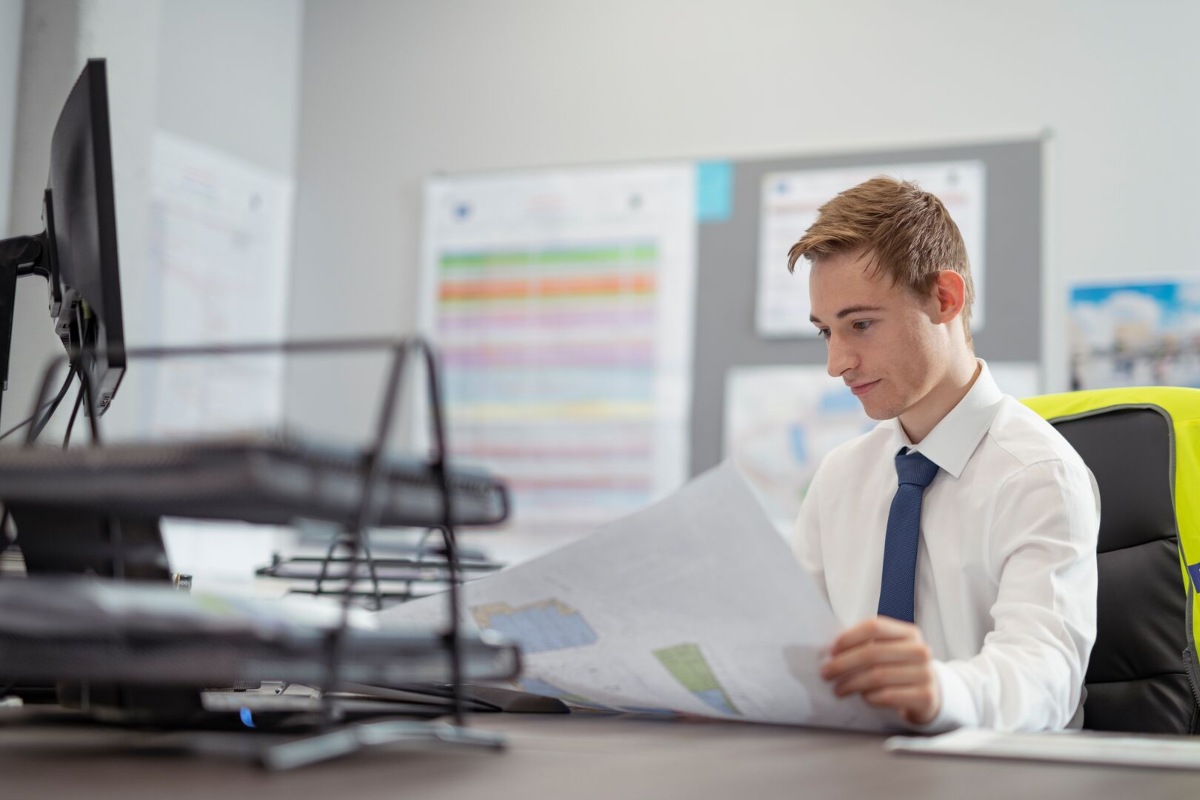 A man sitting at a desk in an office. He is reviewing a plan on a large piece of paper.