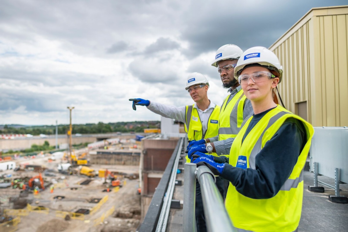 Three students standing on a balcony surveying a building site. They are wearing hi vis and hard hats. 