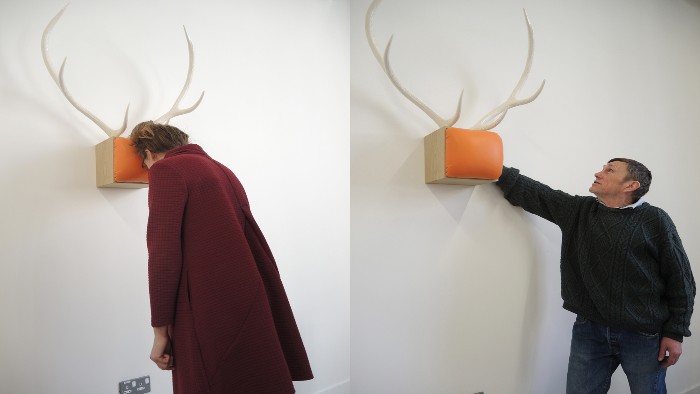 A piece from the domestication project. A two sets of antlers are mounted on the wall with an orange rectangular cushion attached. A man is looking at one and a woman is resting her forehead on another