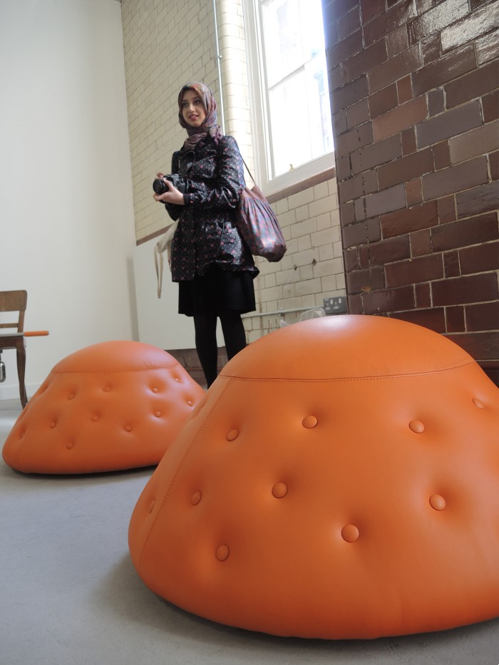 A piece from the Domestication project. Two orange, leather, buttoned, mound-shaped pieces of furniture on the ground with a woman standing behind them 