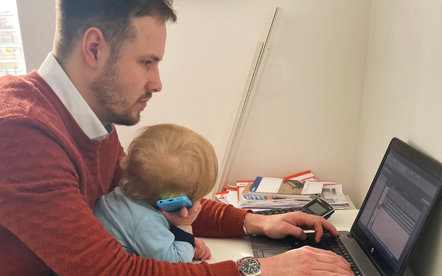 A man working at a laptop in an orange jumper and shirt with a small child on his lap pretending to be on the phone.