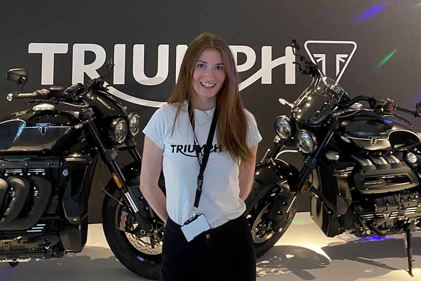 A student stood smiling in front of 2 motorcycles and a triumph company logo on the background