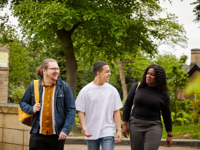 Three students are walking together and chatting on campus, with trees and greenery in the background.