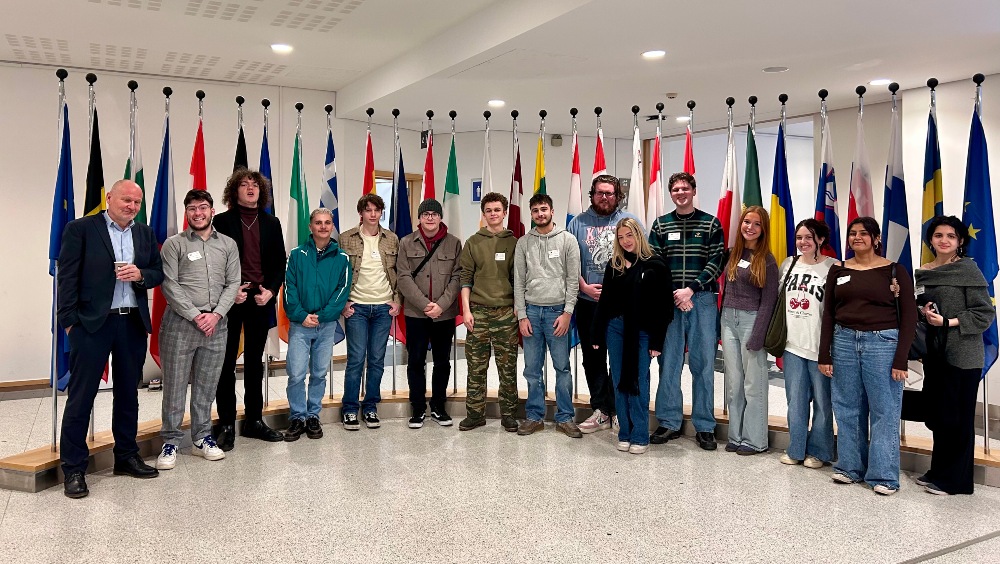 BA (Hons) Politics and International relations students pose for a photo in a grand room with many flags of the EU. 