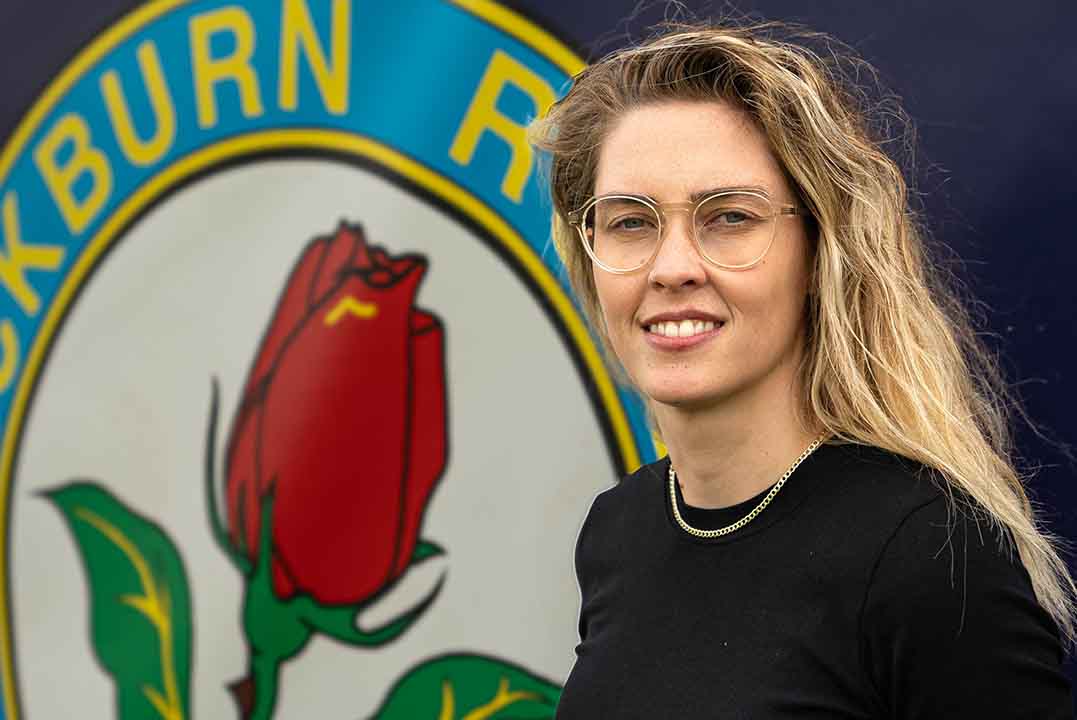 Image of a student with long blonde hair smiling, she wears glasses and stands in front of an image of a rose on the wall behind
