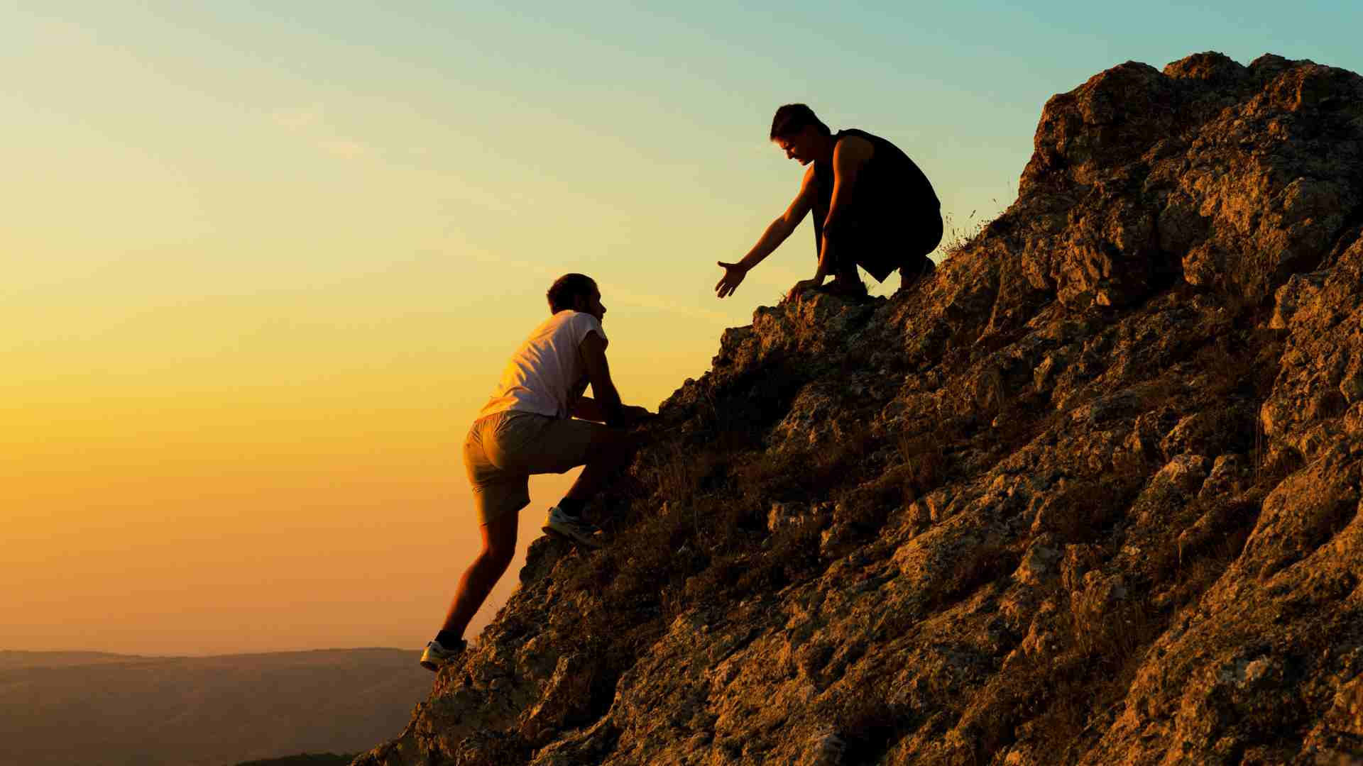 Two people climbing a mountain. One reaches back to help the other climb up. 