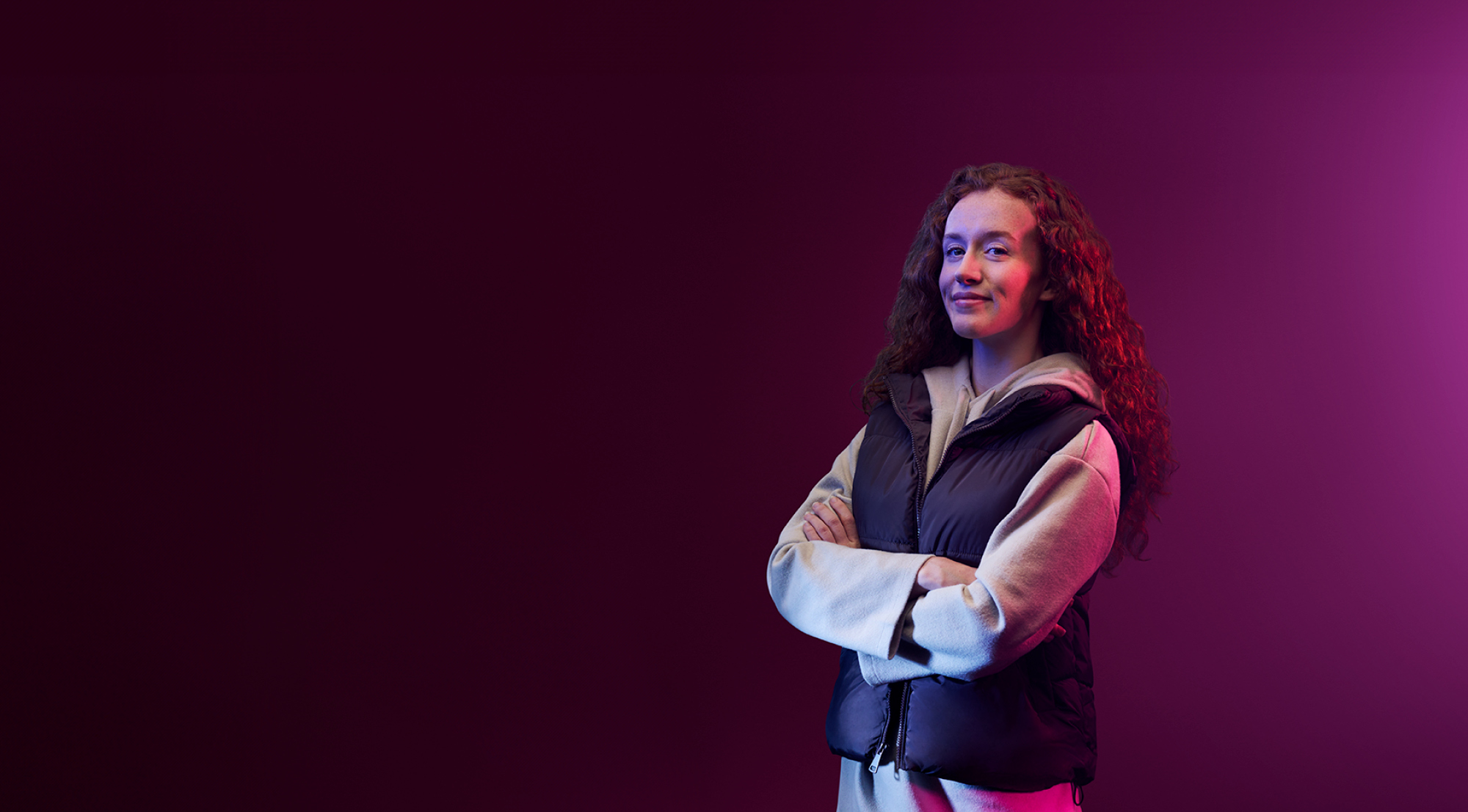 Purple background with student in a body warmer over a hoody smiling with their arms folded