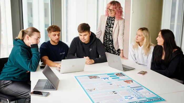 A group of students sat in a classroom around a table in conversation. An academic is stood observing and laptops are on the table in front of them.