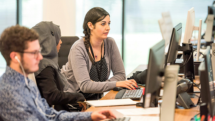 Three students are working on desktop computers. One of them is wearing headphones. The other two may be working together.